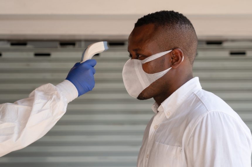 health control staff in virus protection clothing and face shield scanning temperature with digital device to african american businessman to check for fever in coronavirus covid-19 spreading prevention